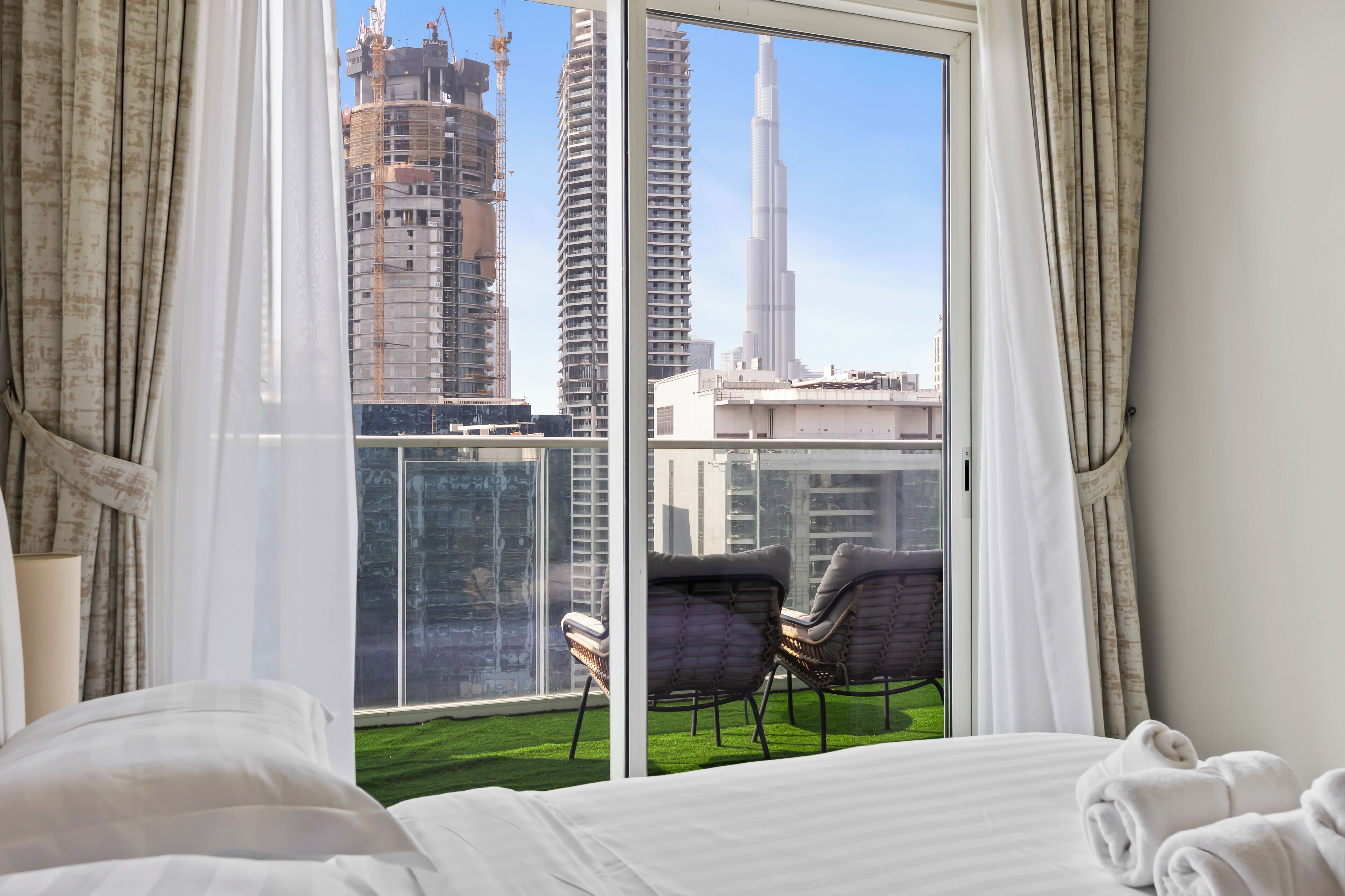 A hotel room with a large window offering a view of high-rise buildings and a balcony with chairs.