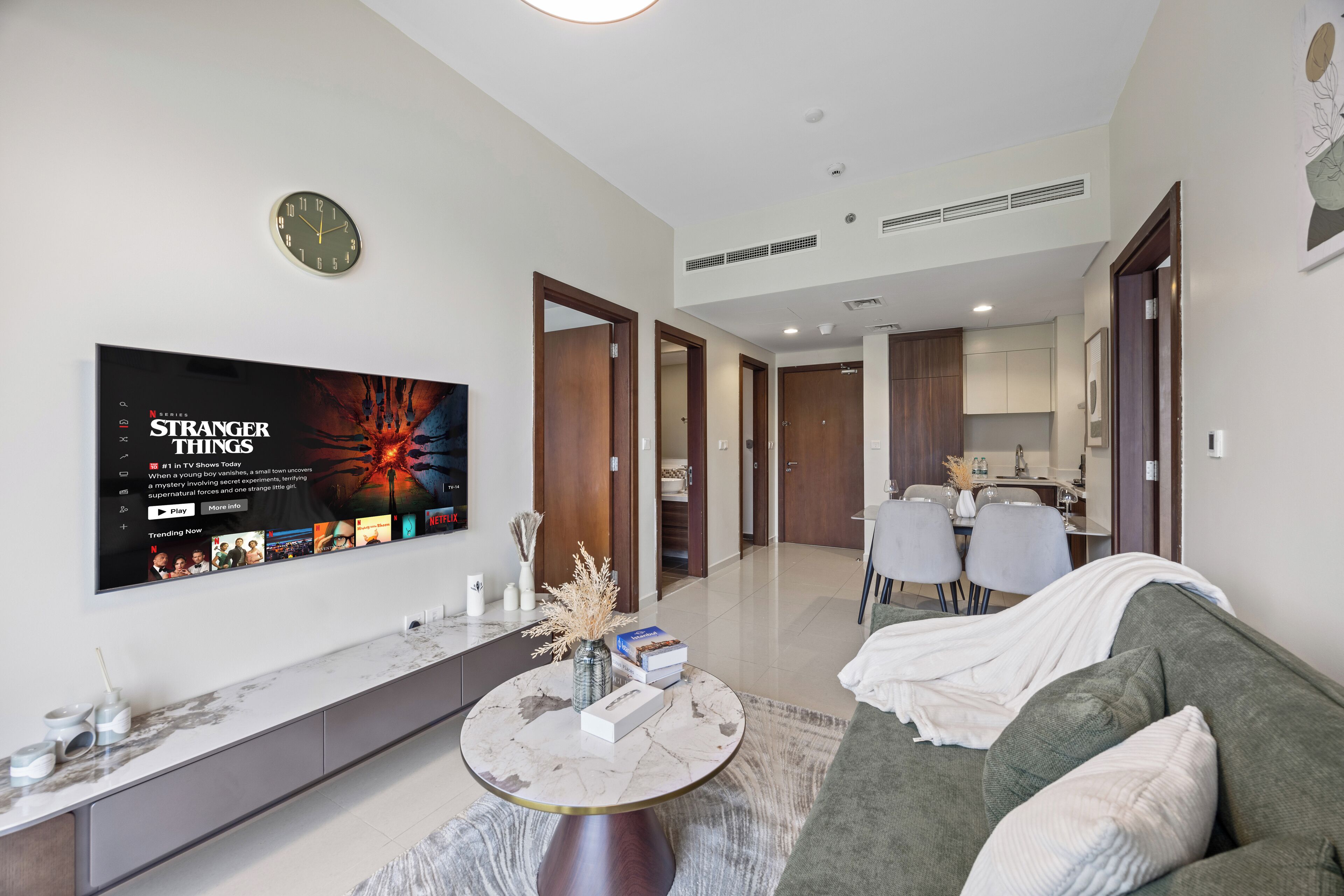 A modern living room with a flat-screen TV displaying 'Stranger Things,' a marble coffee table, a grey sofa, and a dining area in the background.