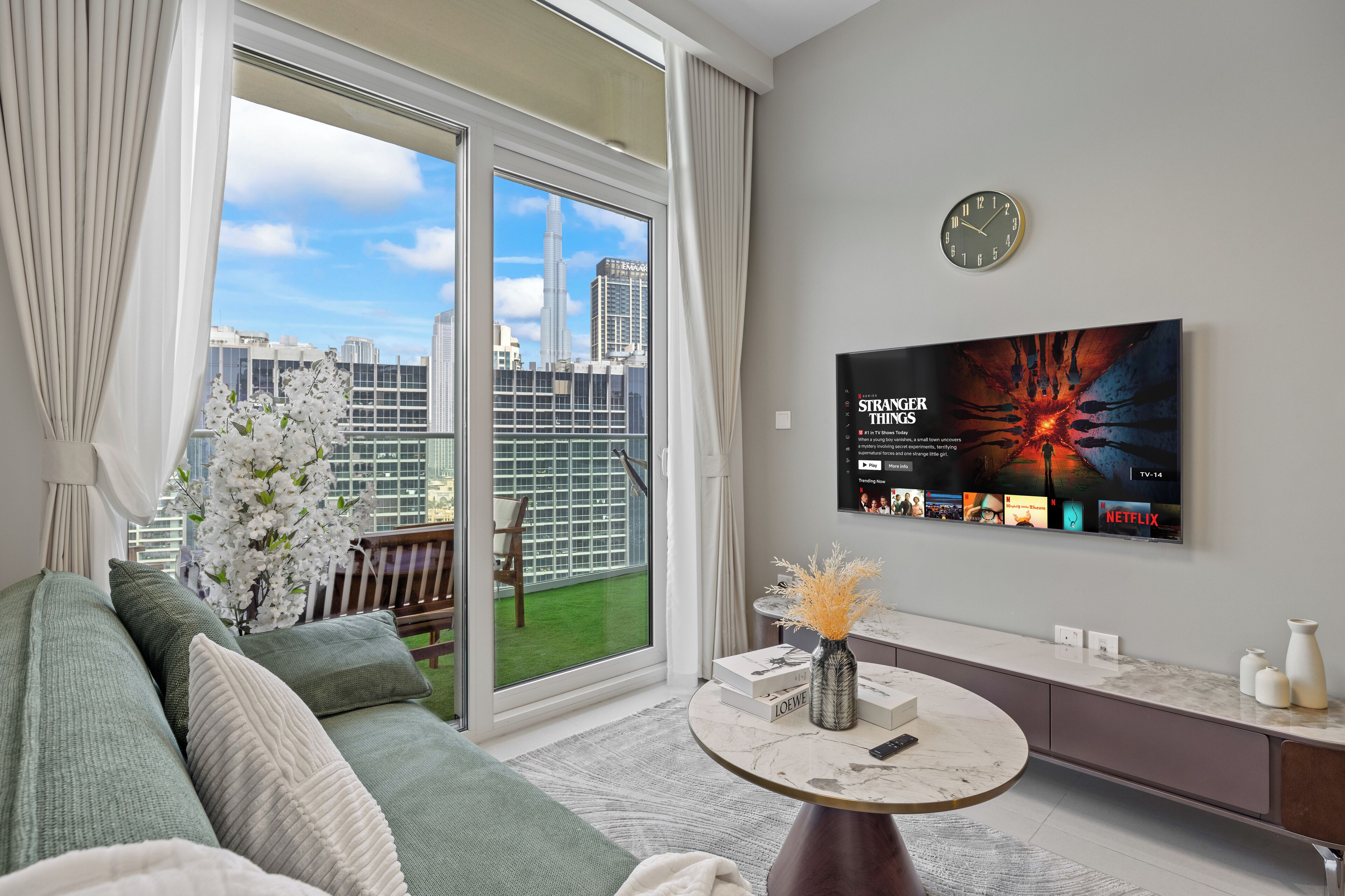 A modern living room with a large TV, a sofa, a coffee table, and a view of a cityscape through a sliding glass door.
