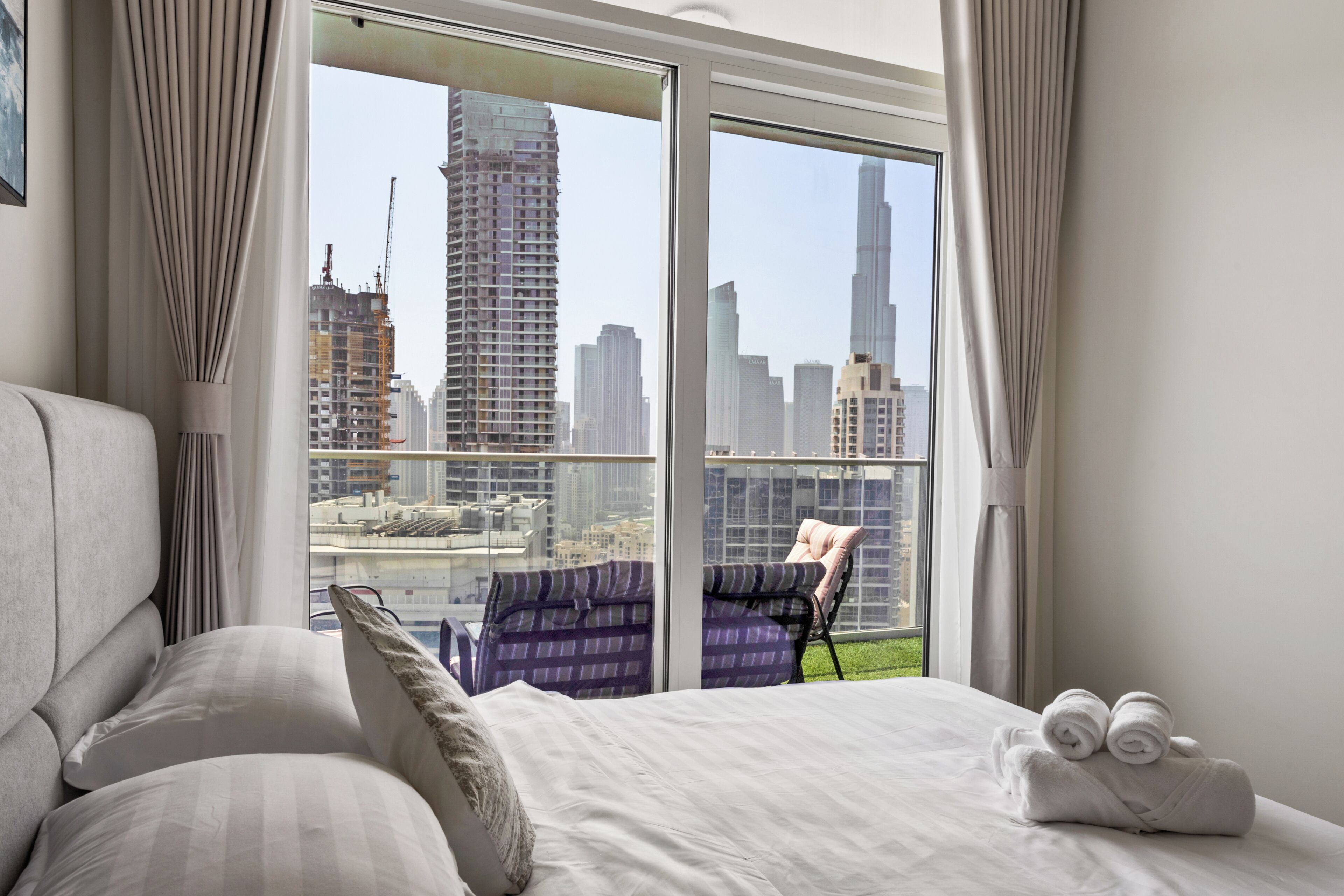 A hotel room with a large window offering a city view, a bed with white linens, a chair with a cushion, and a pair of rolled towels on the bed.