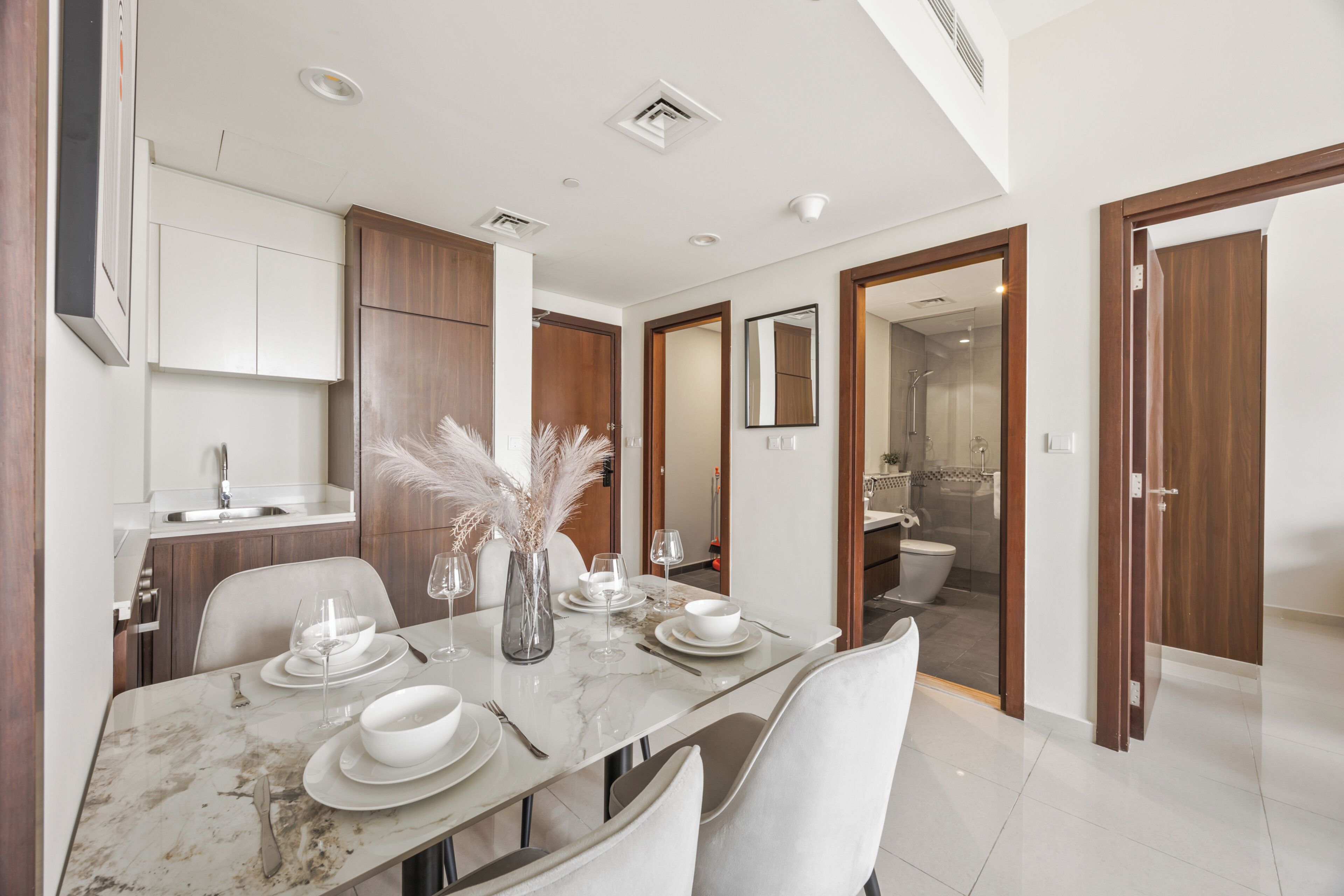 A modern dining area with a marble table, white plates, and chairs. There is a kitchen area with wooden cabinets and a sink.