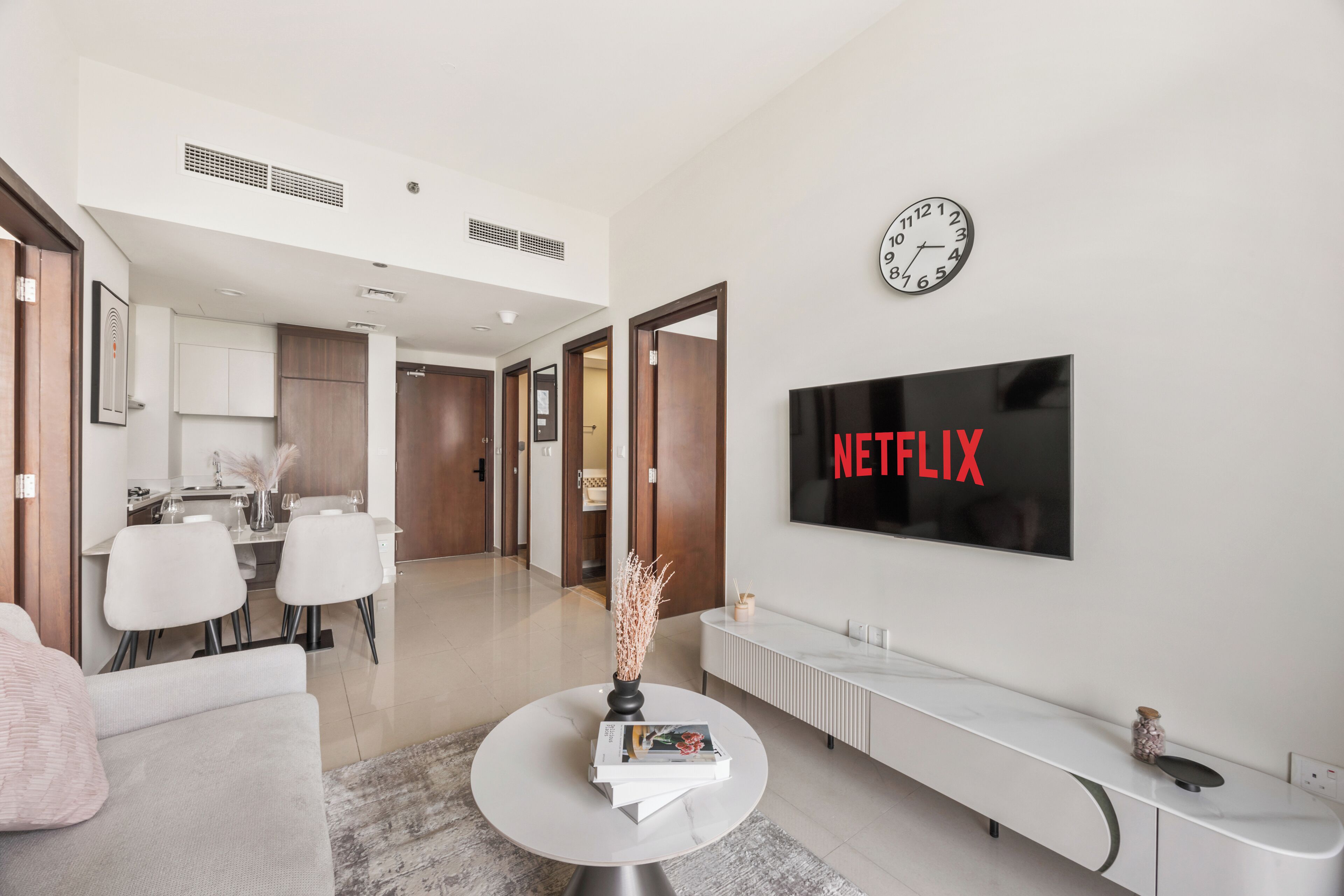 Ein modernes Wohnzimmer mit einem weißen Sofa, einem runden Couchtisch mit Büchern und einer Vase, einem weißen Konsolentisch und einem an der Wand montierten Fernseher, der das Netflix-Logo zeigt.