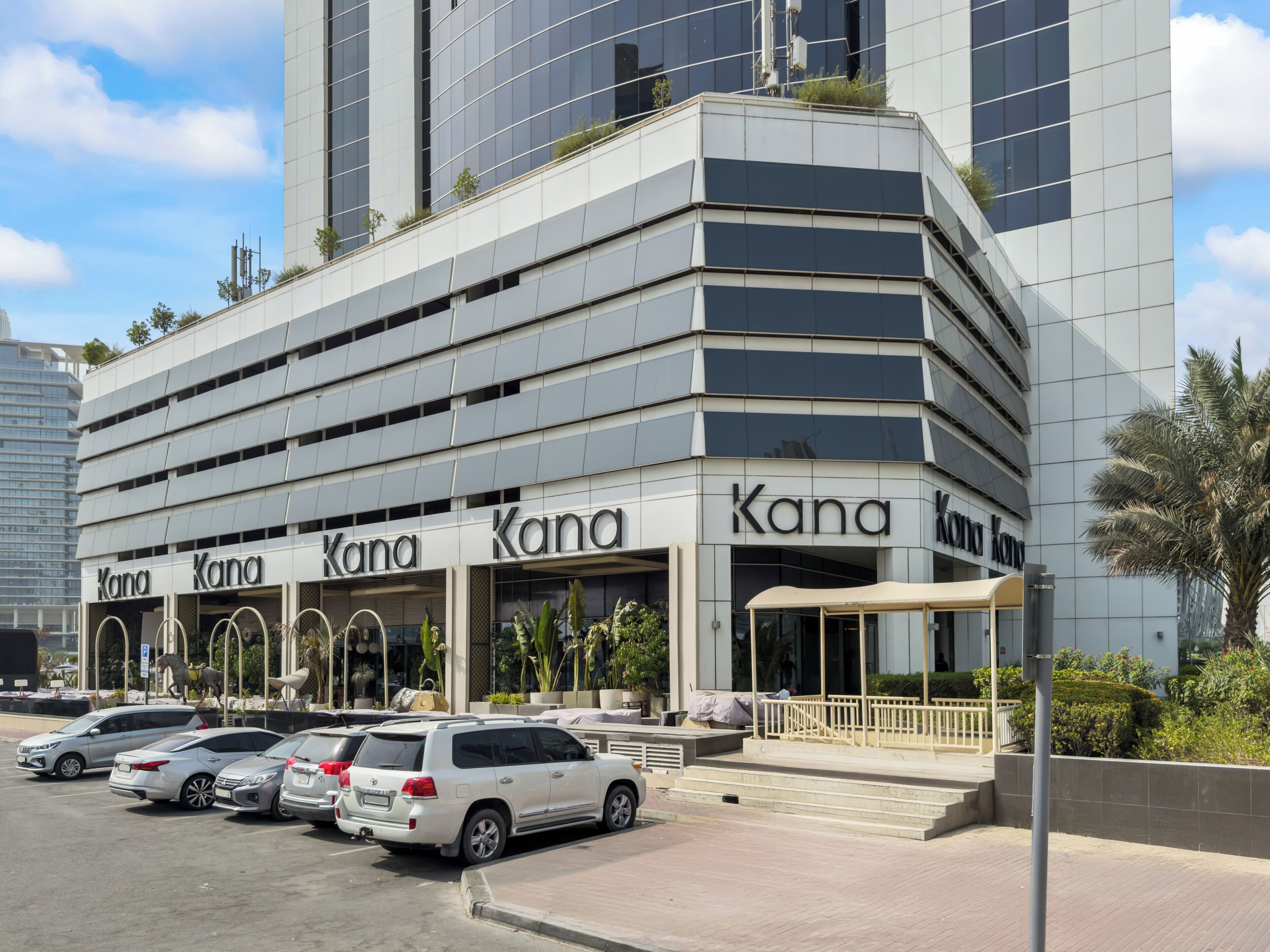 A modern building with 'Kana' signage, featuring a parking area with several cars, a covered entrance, and a staircase leading to the entrance.