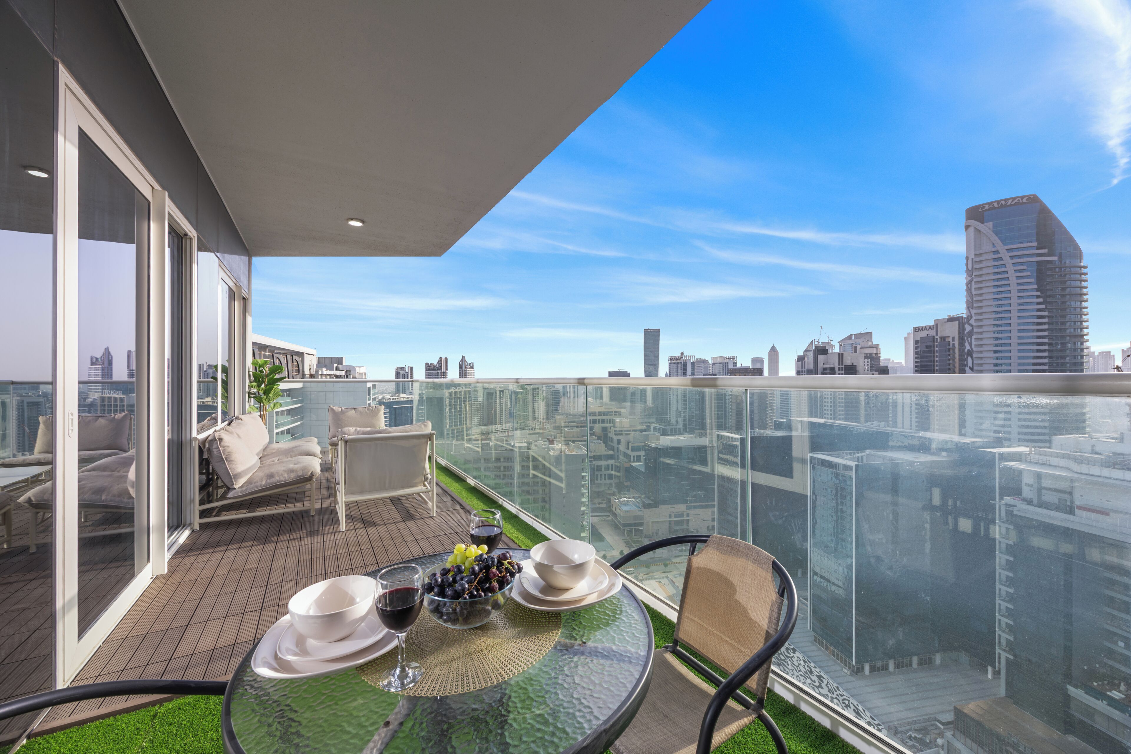 A modern balcony with a glass railing, furnished with a table, chairs, and a small garden area.