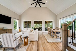 A spacious living room with a high ceiling, a wooden staircase, and large windows offering views of the outdoors.