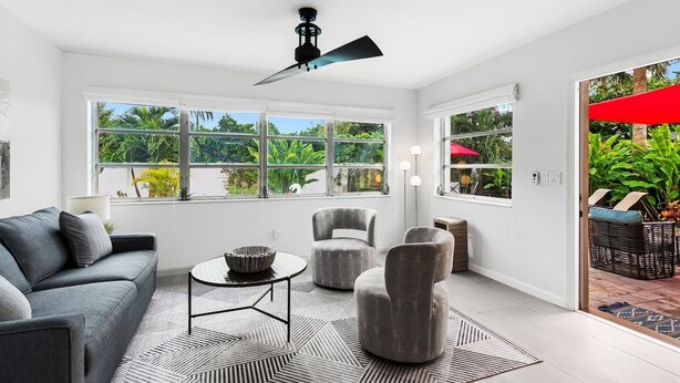 A modern living room with a grey sofa, round coffee table, and grey armchairs. Large windows offer views of greenery. - Oasis Backyard with Heated Pool! | Oakland Park (Wilton Manors)