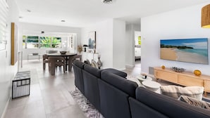 A modern living room with a dark sofa, a dining area with a table and chairs, and a large TV on the wall. - Oasis Backyard with Heated Pool! | Oakland Park (Wilton Manors)