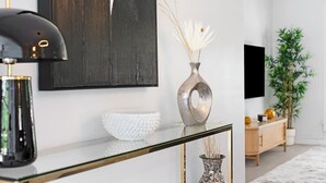 A modern living room with a glass coffee table, a decorative vase with dried plants, a white textured bowl, a black abstract painting, a wooden cabinet, and a large potted plant. - Oasis Backyard with Heated Pool! | Oakland Park (Wilton Manors)