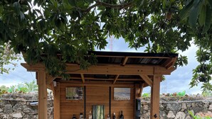 A wooden outdoor bar area with a stone wall backdrop, three bar stools, and a wooden pergola providing shade. - appartement,calme,securise entre mer et campagne (Juan Dolio)