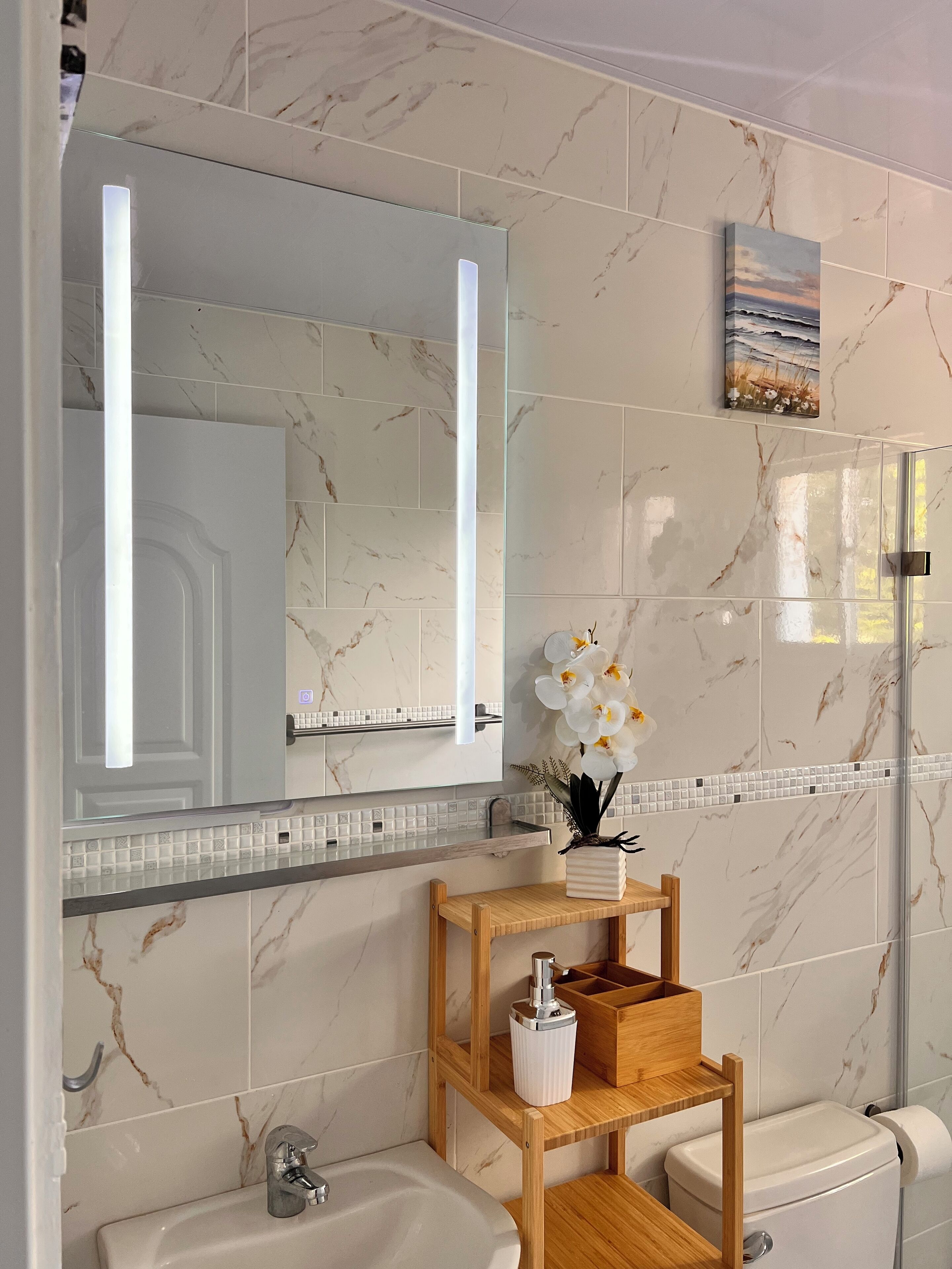 A bathroom with marble tiles, a large mirror, a wooden vanity, a toilet, and a small shelf with a vase of flowers.
