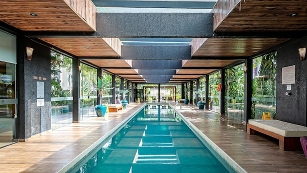 A modern indoor pool area with a long, narrow pool, wooden ceiling beams, and glass walls offering views of greenery outside. - Apartment in Thai Beach Home Spa Tbs1105 (Florianópolis)