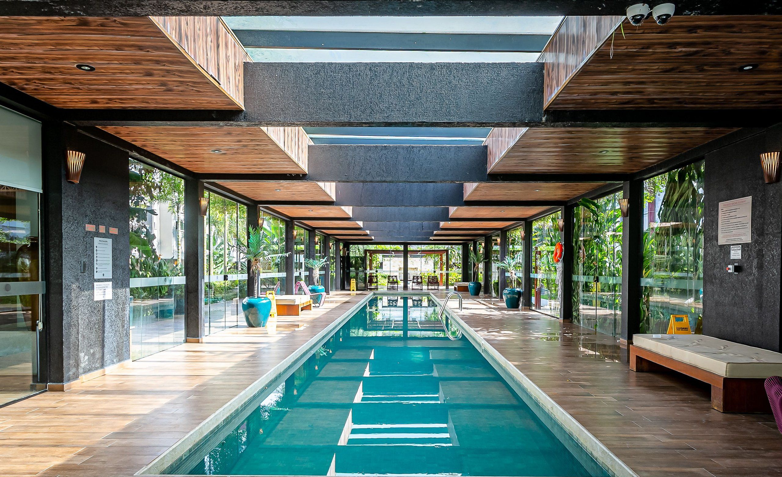 A modern indoor pool area with a long, narrow pool, wooden ceiling beams, and glass walls offering views of greenery outside.