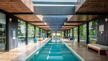 Un área de piscina interior moderna con una piscina larga y estrecha, vigas de techo de madera y paredes de vidrio que ofrecen vistas a la vegetación exterior.