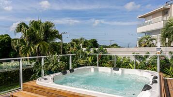 Un jacuzzi en una terraza de madera con vistas a un edificio moderno y abundante vegetación.