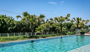 A swimming pool with clear blue water, surrounded by lush green palm trees and a fence. - Apart in Thai Beach Home Spa Cond sea Front Tbs106 (Florianópolis)