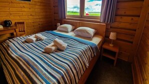 A wooden cabin bedroom with a bed, pillows, a nightstand, a lamp, and a view of the outdoors. - Double room with breakfast in Świnoujście (Świnoujście)