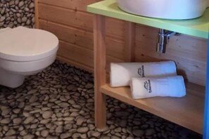 A wooden cabin-style bathroom with a toilet, a shelf with towels, and a stone floor.