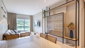 A modern living room with a glass dining table, a sofa with cushions, a TV mounted on the wall, and a decorative shelf with glassware and a vase. - Modern 2BR Apt Close to the Beach Stl0702 (Balneário Camboriú)