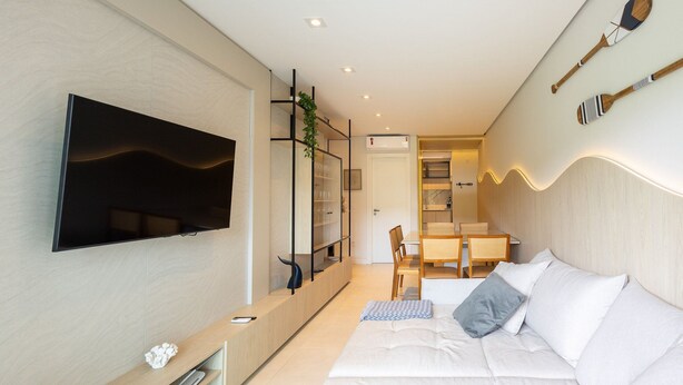 A modern living room with a flat-screen TV, a sofa with cushions, a dining area with a table and chairs, and a kitchen visible through an open door. - Modern 2BR Apt Close to the Beach Stl0702 (Balneário Camboriú)