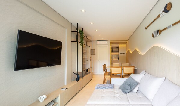 Una sala moderna con una televisión de pantalla plana, un sofá con cojines, una zona de comedor con una mesa y sillas, y una cocina visible a través de un umbral abierto.