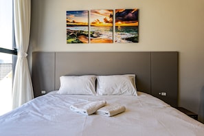 A neatly made bed with white linens and two pillows, a wall-mounted artwork depicting a sunset over the ocean, and a window with curtains.