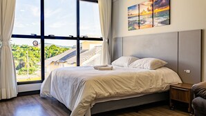 A bedroom with a large window, a bed, a nightstand, and a painting of a sunset over the ocean. - Studio in Condominium With Pool 700m Beach Sti210 (Florianópolis)