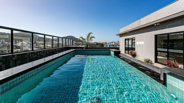 A rooftop infinity pool with clear blue water, surrounded by a modern building with large windows and a view of the cityscape and mountains in the background. - New Studio Well Located 750m From the Beach Sti209 (Florianópolis)