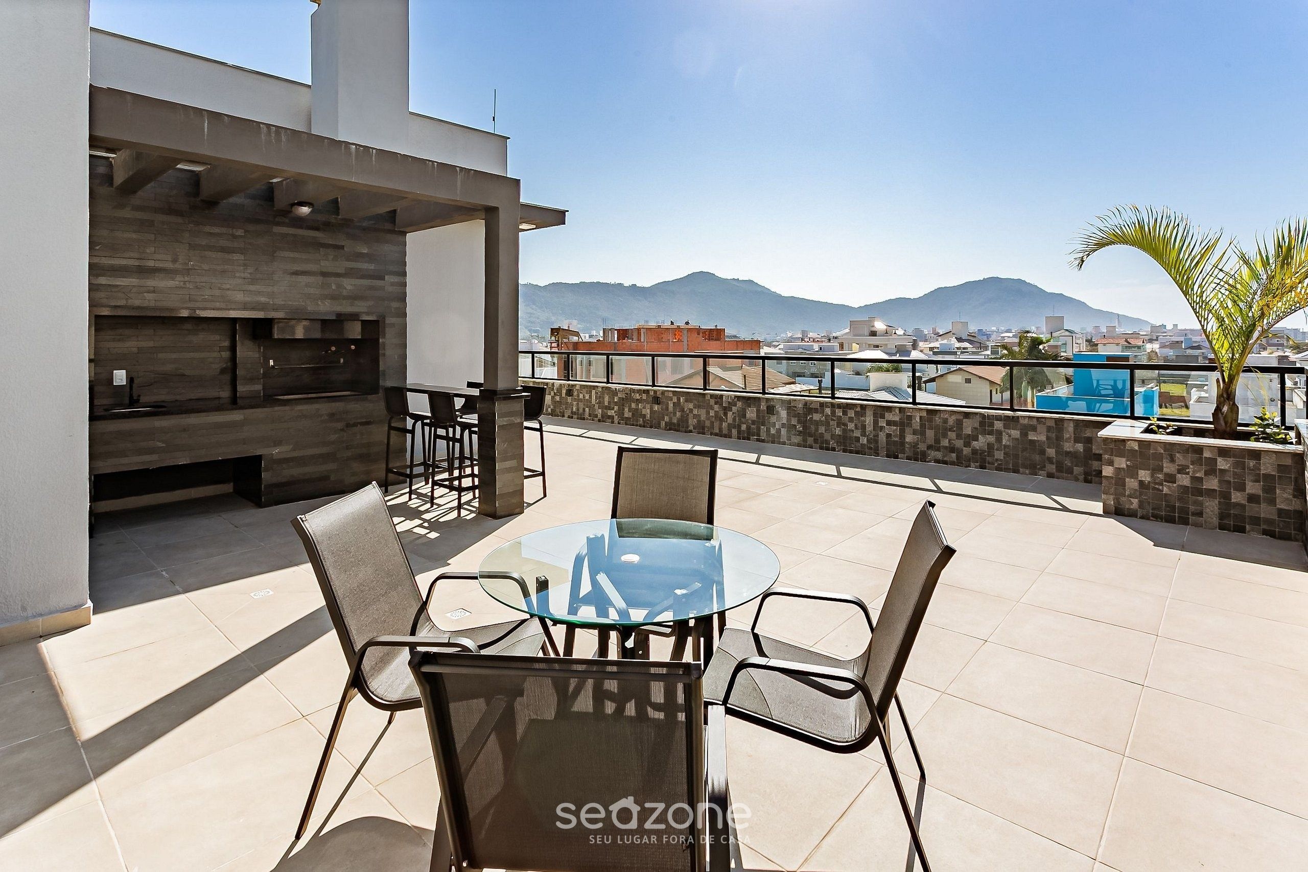An outdoor seating area with a glass table and chairs, a barbecue grill, and a view of a cityscape with mountains in the background.