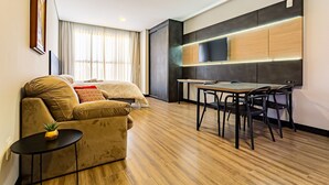 A modern hotel room with a sofa, a small table with a plant, a dining area with a table and chairs, and a large TV mounted on the wall. - New Studio Well Located 750m From the Beach Sti209 (Florianópolis)