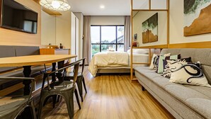 Una habitación de hotel moderna con piso de madera, una cama con ropa blanca, un sofá con cojines decorativos, una mesa de comedor con sillas y un ventanal con vistas al exterior.