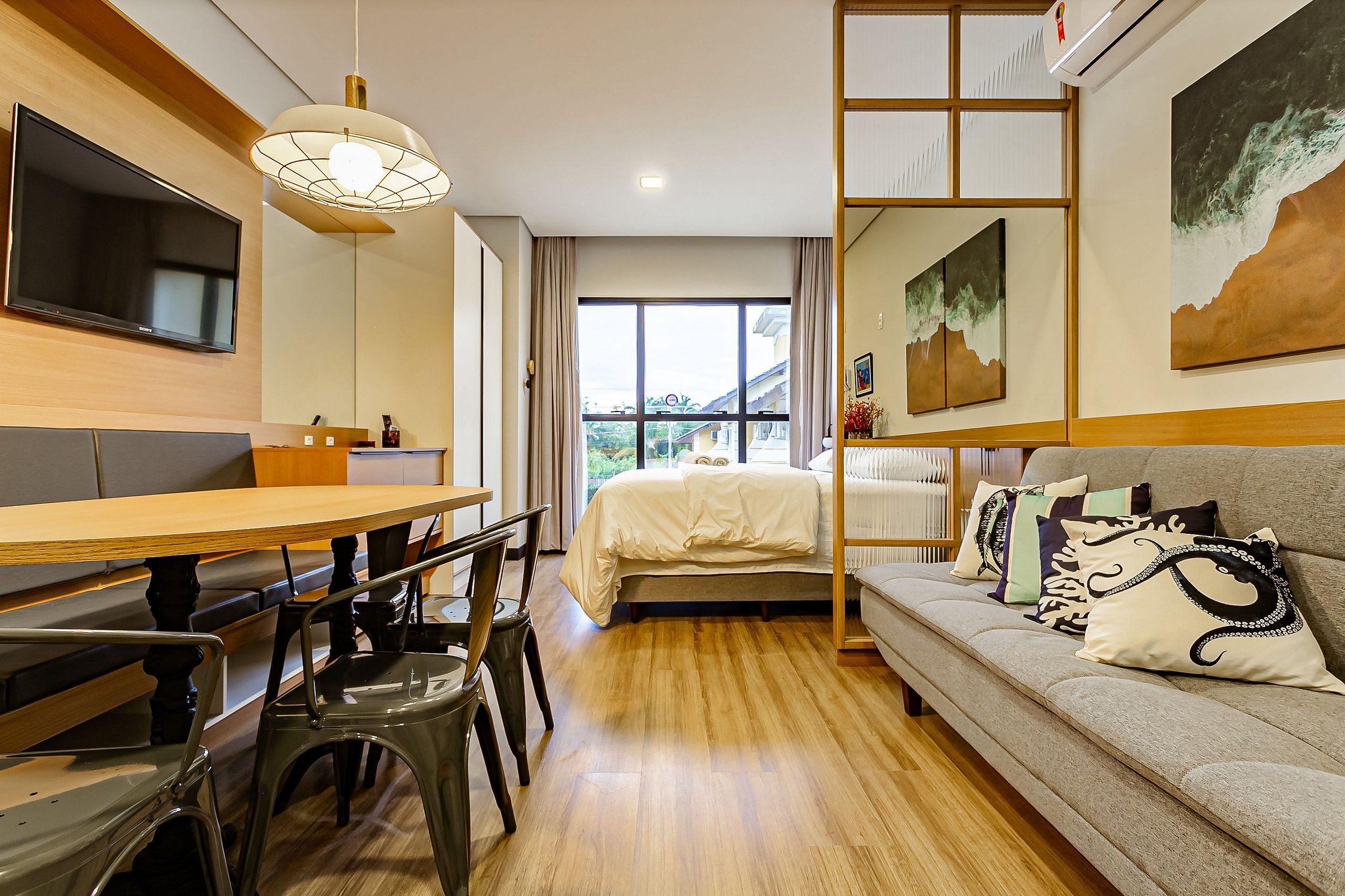 Quarto de hotel moderno com piso de madeira, cama com roupa de cama branca, sofá com almofadas decorativas, mesa de jantar com cadeiras e uma janela grande que oferece vista para o exterior.