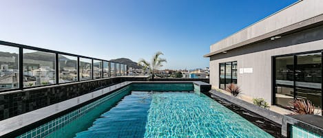 A rooftop swimming pool with clear blue water, surrounded by a modern building with large windows and a view of the cityscape.
