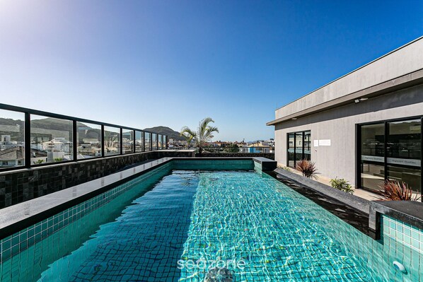 A rooftop swimming pool with clear blue water, surrounded by a modern building with large windows and a view of the cityscape.
