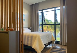 A bedroom with a bed, a window with curtains, a view of greenery outside, and a wooden partition.