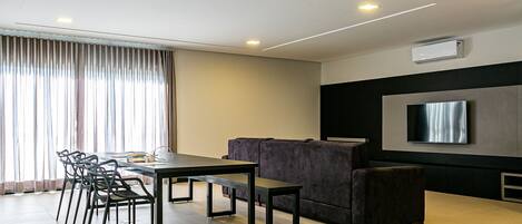 Un salon moderne comprenant un coin repas, un téléviseur à écran plat fixé au mur et une grande fenêtre avec des rideaux.