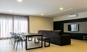 A modern living room with a dining area, a flat-screen TV mounted on the wall, and a large window with curtains.