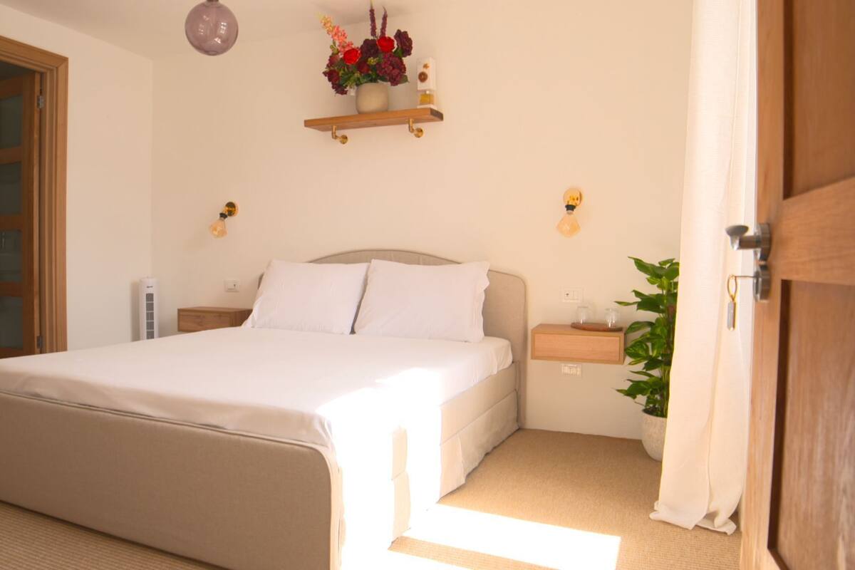 A bedroom with a bed, bedside tables, a nightstand, a wall-mounted shelf with flowers, and a door.