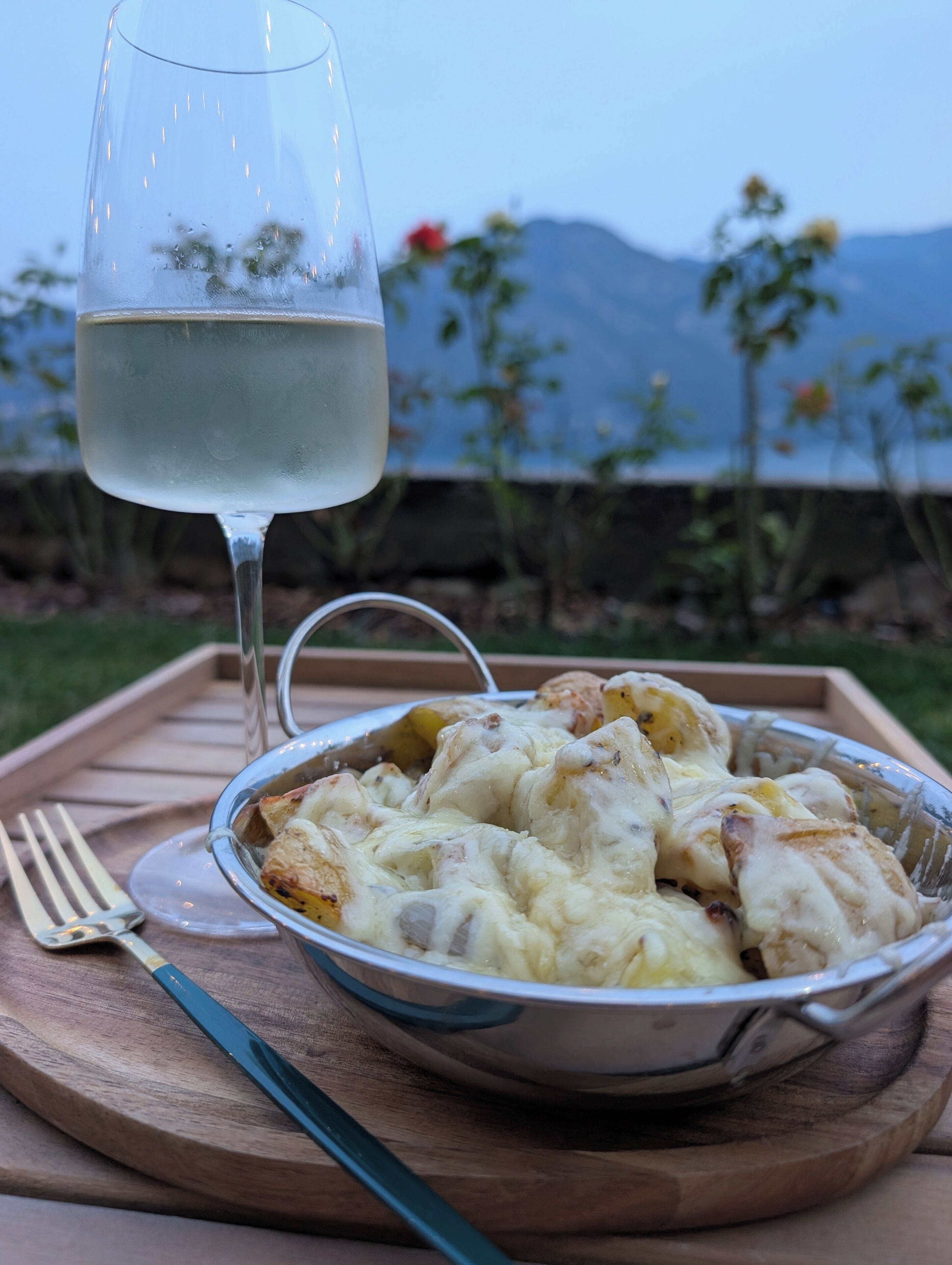 A bowl of food on a wooden tray with a fork and a glass of white wine.