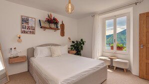 A bedroom with a large bed, bedside tables, a bedside lamp, a window with a view of mountains, and a wooden door. - La Casa Della Seta  (Tremezzo)