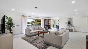 A spacious living room with a sofa set, a coffee table, and a TV. There is a large window with curtains, a plant, and a balcony visible.
