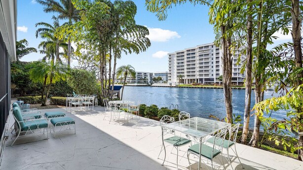 A poolside area with lounge chairs and tables, surrounded by palm trees and a view of a building across the water.