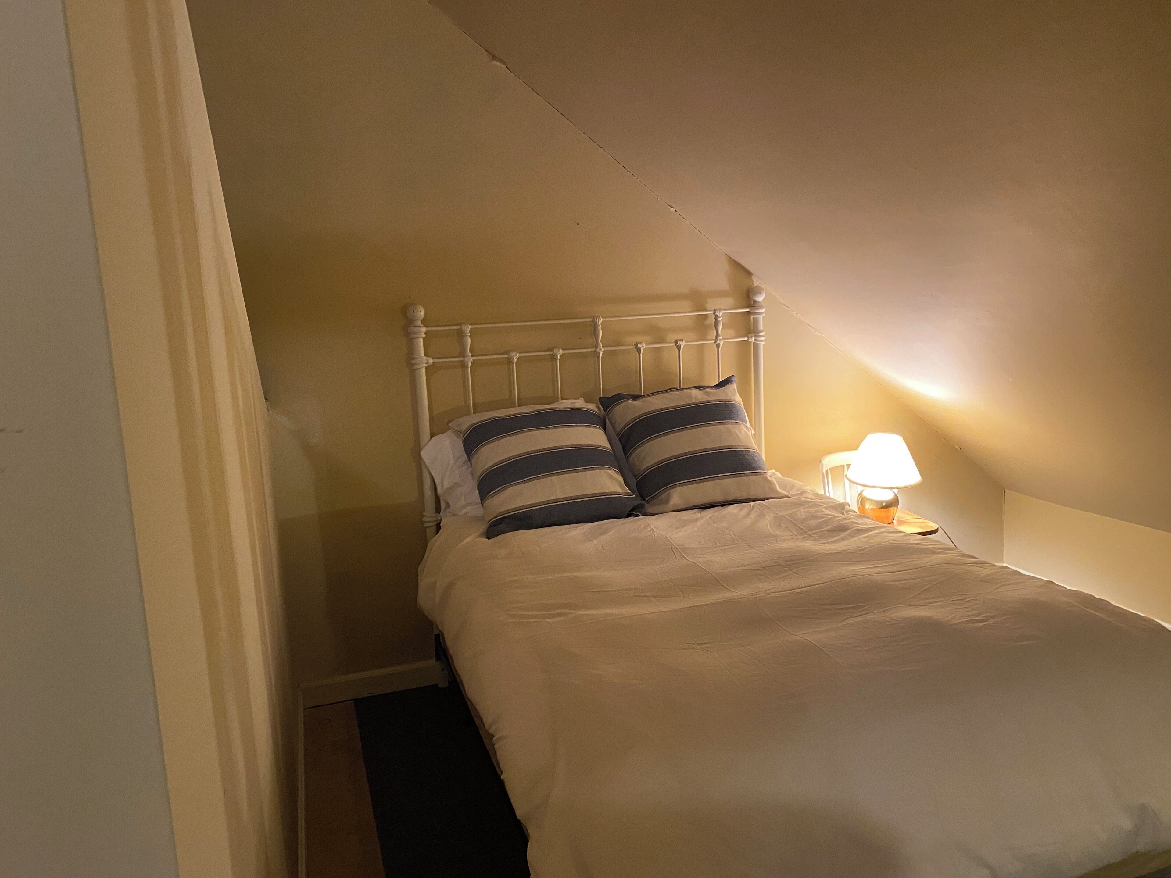 Una habitación pequeña y estrecha con una cama individual, una mesita de noche con lámpara y techo inclinado.