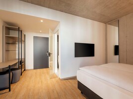 Un dormitorio moderno con una cama, televisión montada en la pared, un escritorio con silla y un armario.