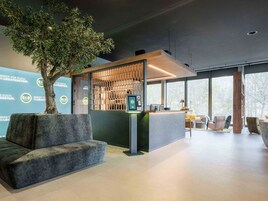 Un área de recepción moderna con mostrador, zona de estar y un amplio ventanal con vistas a árboles.