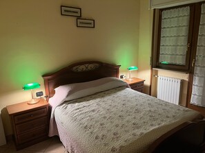 A bedroom with a bed, two bedside tables, a nightstand, a window with lace curtains, and a wall-mounted light fixture.