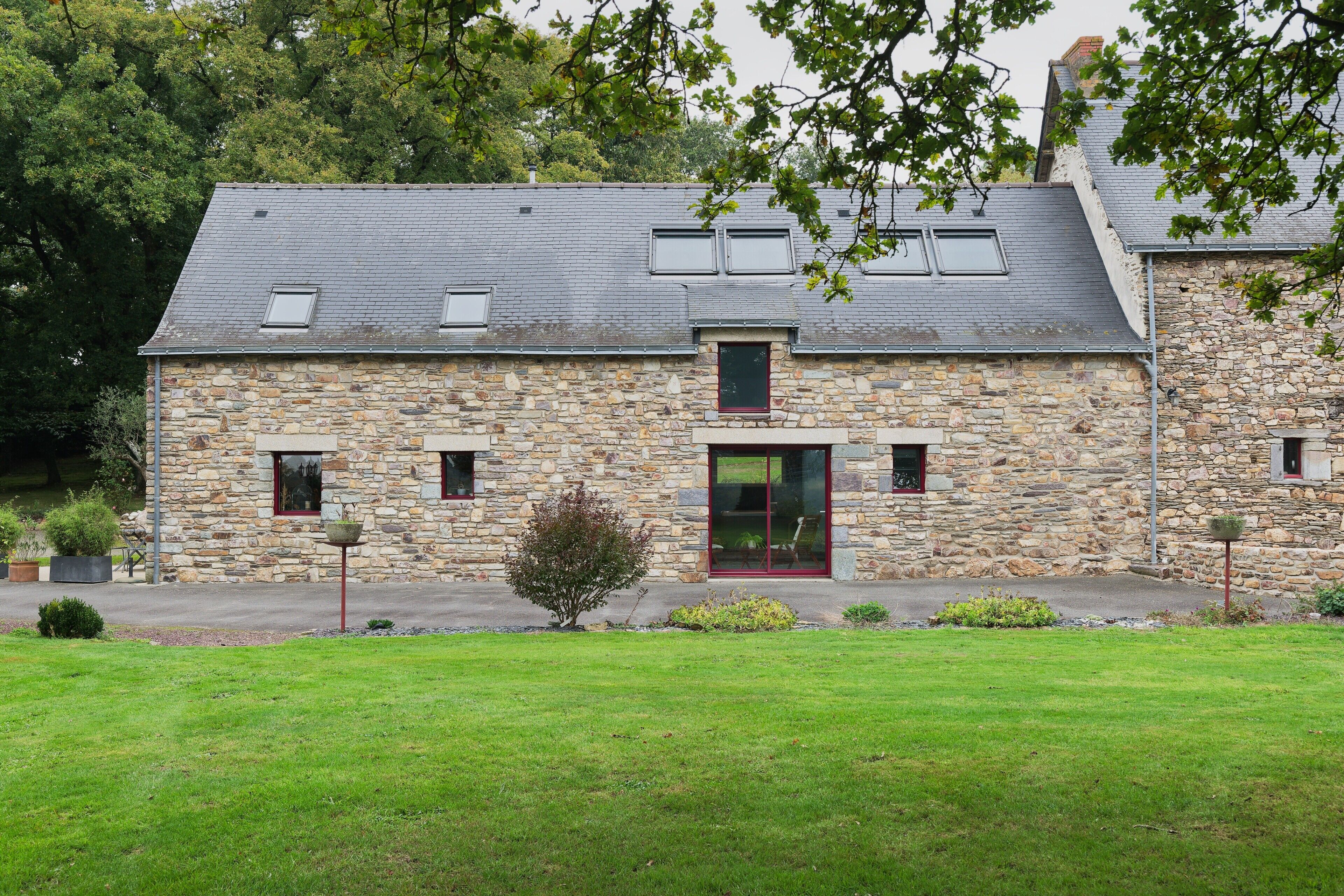 Un bâtiment en pierre doté d’un toit en pente, une entrée centrale avec une porte rouge et un petit jardin à l’avant.