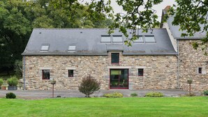 A stone building with a sloped roof, a central entrance with a red door, and a small garden area in front. - Holiday Home 'La Clémençaie' with Private Terrace, Garden and Wi-Fi (Réminiac)