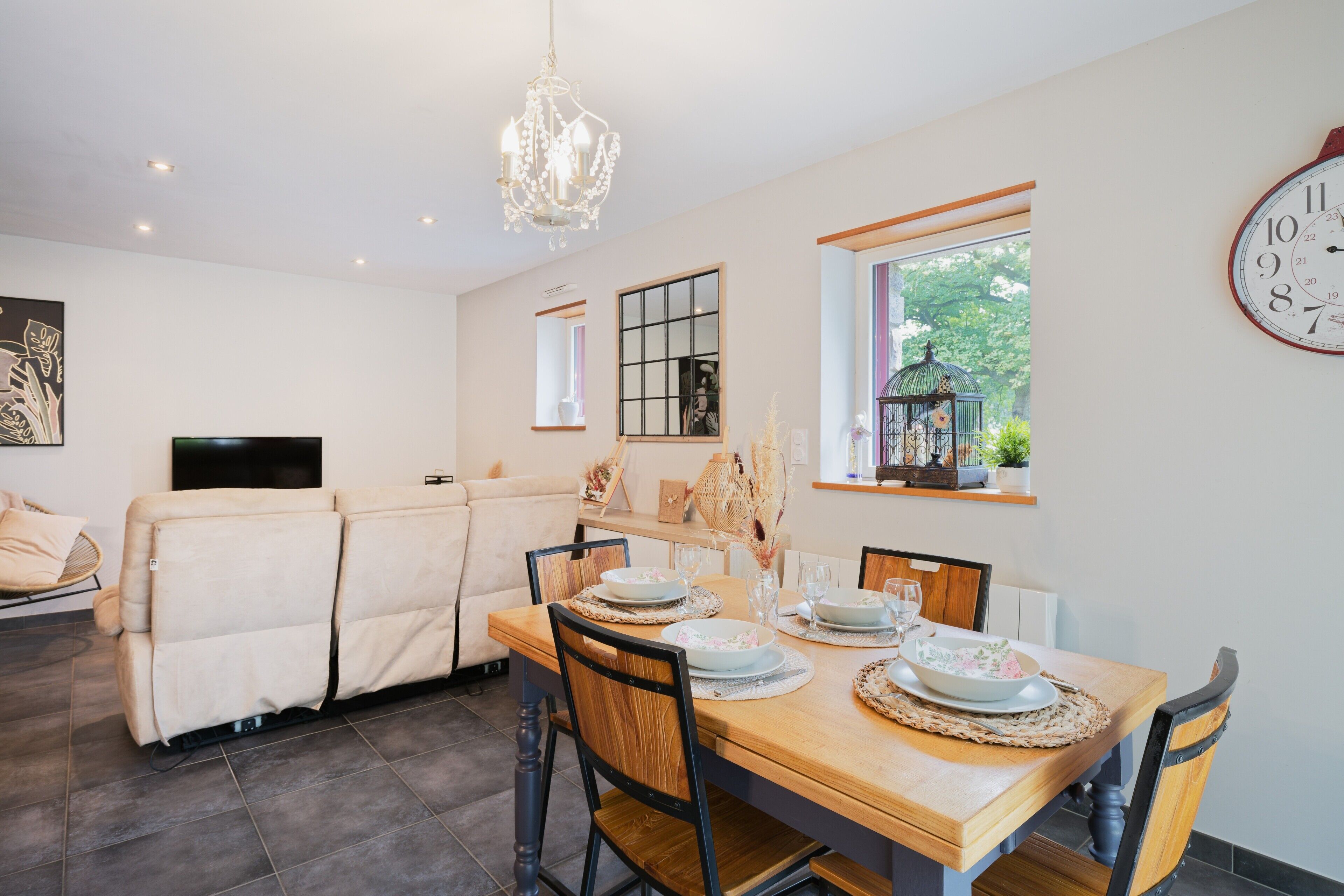 Un coin repas avec une table en bois dressée pour quatre personnes, un canapé, un téléviseur et une horloge au mur.
