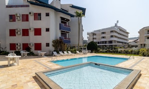 A swimming pool with lounge chairs and tables, surrounded by buildings with balconies and red shutters. - Comfortable Apart 150m From Praia Brava Rra0205 (Florianópolis)