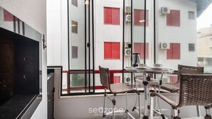 A balcony with a table and chairs, a view of a building with red shutters, and an air conditioning unit. - Apt W/bbq and Access to Praia Brava Rra0103 (Florianópolis)